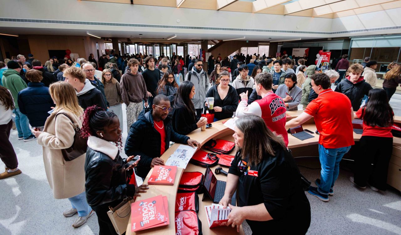 A large group of people mingle and walk around a sunny atrium at a university information fair.