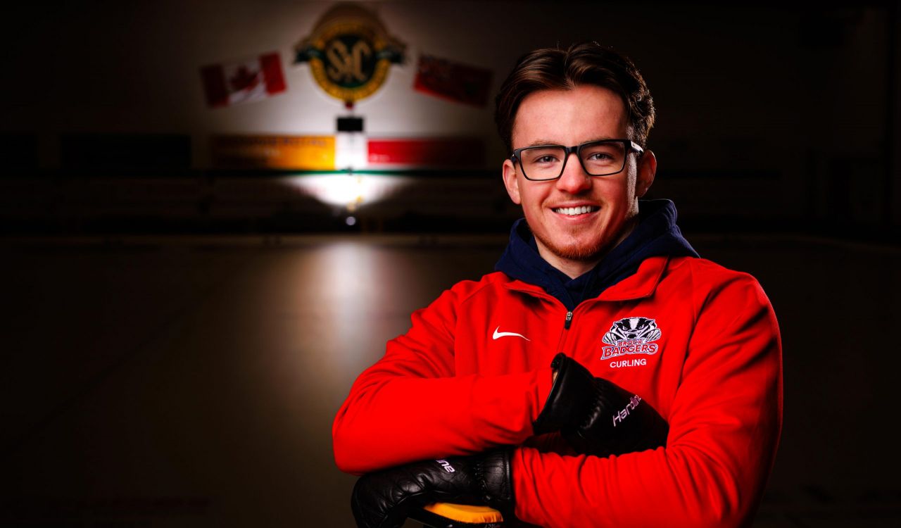 Young male curler in glasses and a red Brock Badgers curling jacket smiles while leaning on his broom on a dimly lit ice rink.