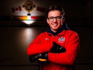 Young male curler in glasses and a red Brock Badgers curling jacket smiles while leaning on his broom on a dimly lit ice rink.