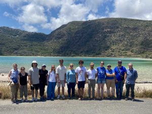 A group of university students stand together in front of a lake with mountains and an archaeological excavation site in the background.