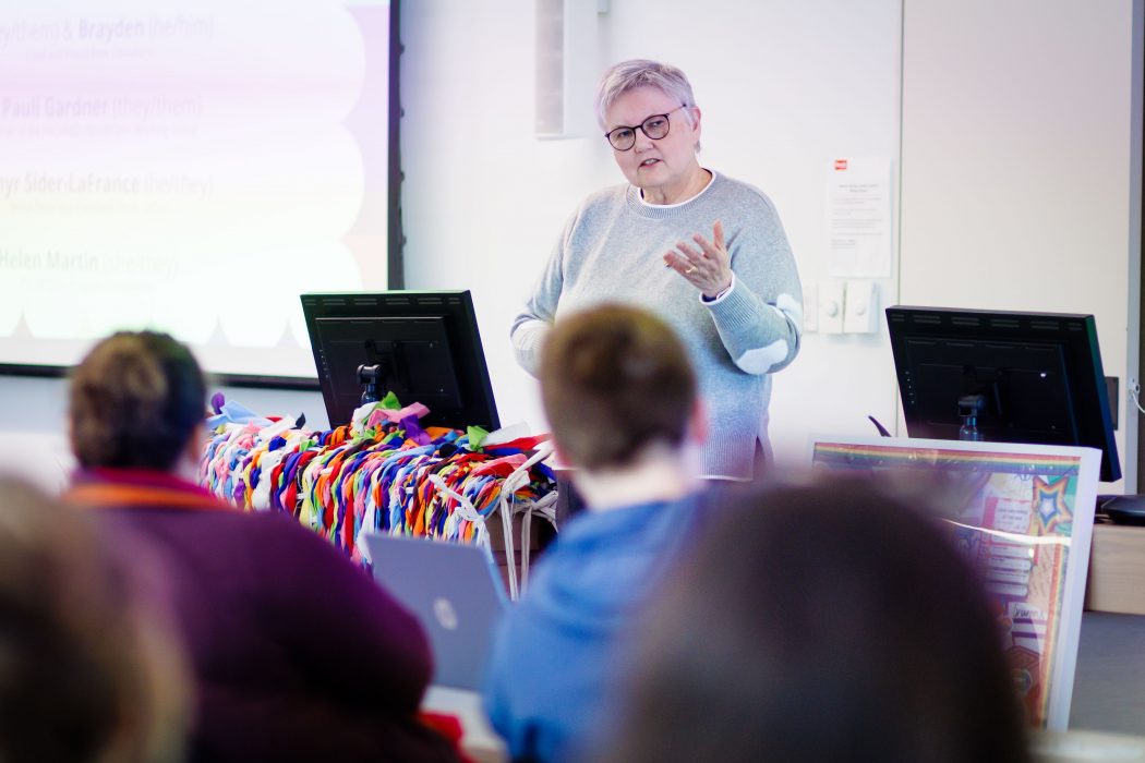 A speaker with short gray hair and glasses gestures while presenting at the front of a classroom