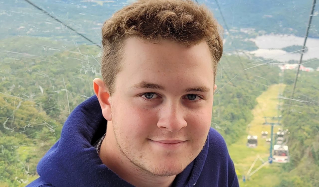 A man wearing a Brock University hoodie smiles while sitting in a cable car traversing a mountain.