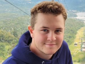 A man wearing a Brock University hoodie smiles while sitting in a cable car traversing a mountain.