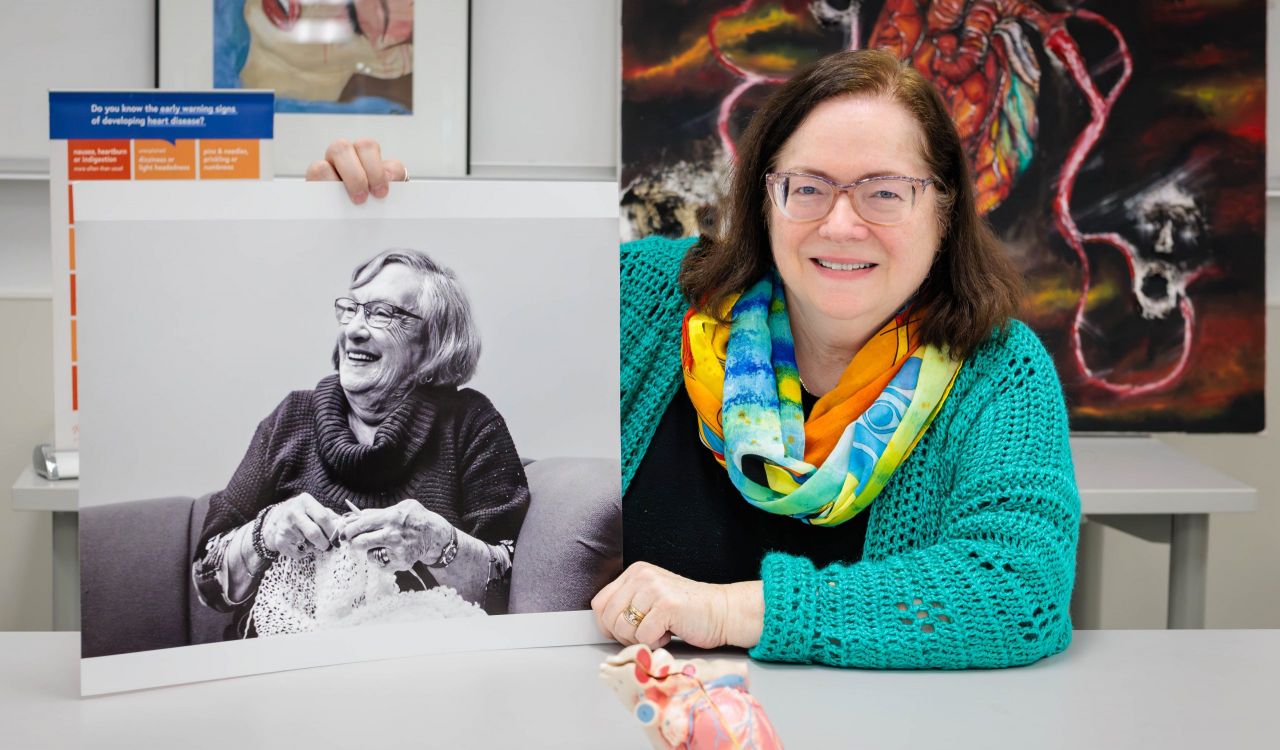 A smiling woman sits at a table holding up a black-and-while photograph of an older woman drinking tea and smiling.