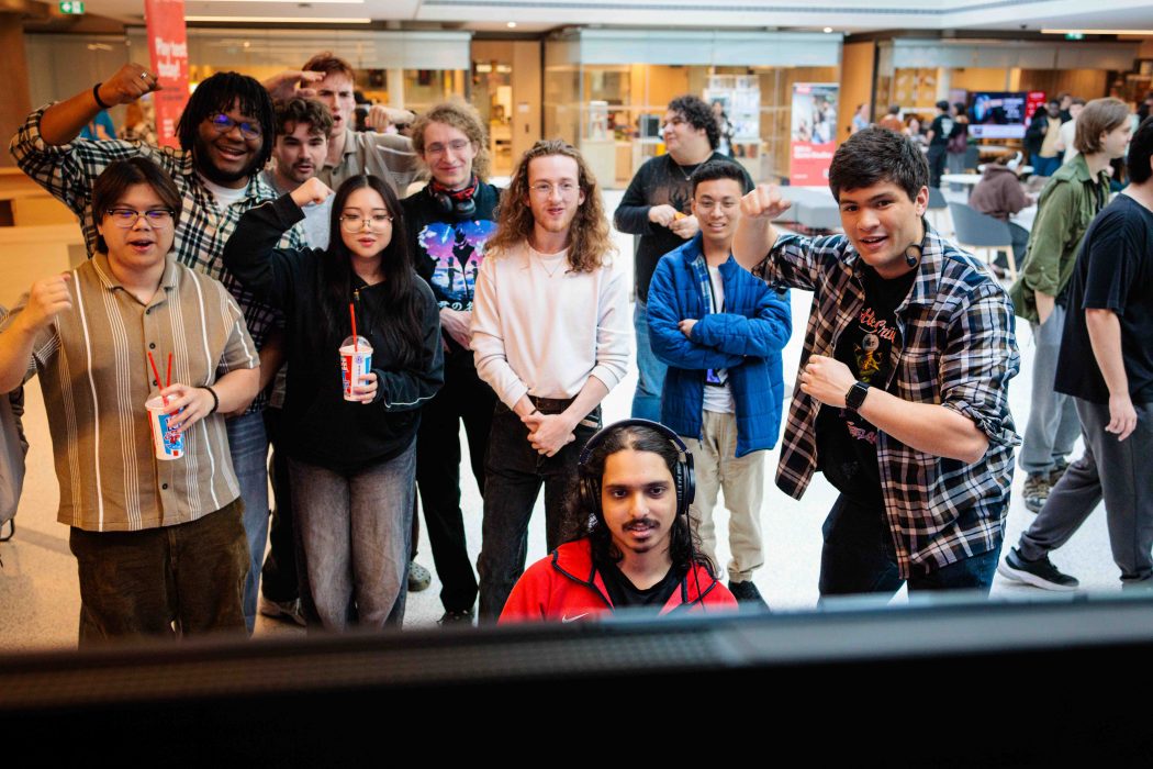 A group of students gathers around a gaming station in a campus atrium, cheering and raising their fists while one player wearing headphones sits in the foreground focused on the screen.