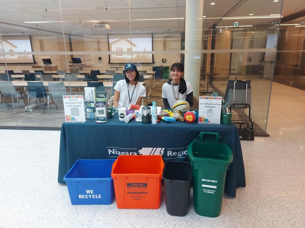 Two people stand behind a table with recycling bins around them at an information fair.