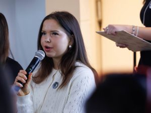 A young woman speaks into a microphone while addressing an audience.