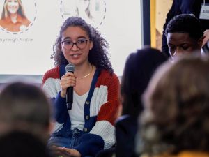 A young woman speaks into a microphone while addressing an audience.