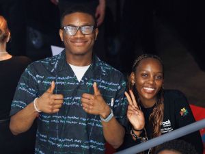 Two people smile for a photo while giving a thumbs up and holding up a peace sign.