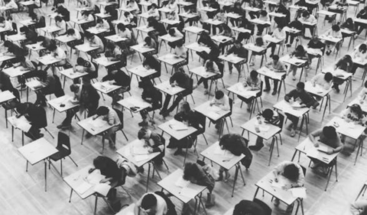 A black and white photo of rows of desks in a gymnasium with students sitting at them taking their exams.