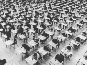 A black and white photo of rows of desks in a gymnasium with students sitting at them taking their exams.
