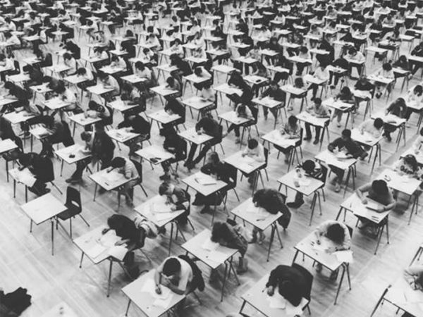 A black and white photo of rows of desks in a gymnasium with students sitting at them taking their exams.