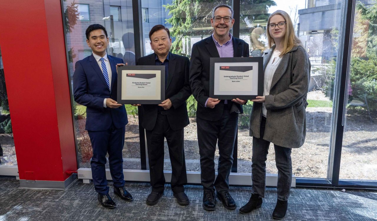 The photo has two people in the center who are both receiving plaques from students standing on the outside. The plaque reads Student-Voted Teaching Award.