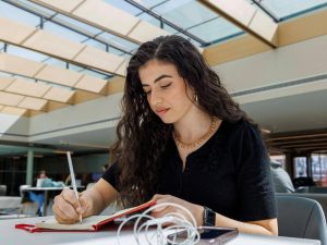 A young woman sits at a table writing notes in a notebook in a sunny room.