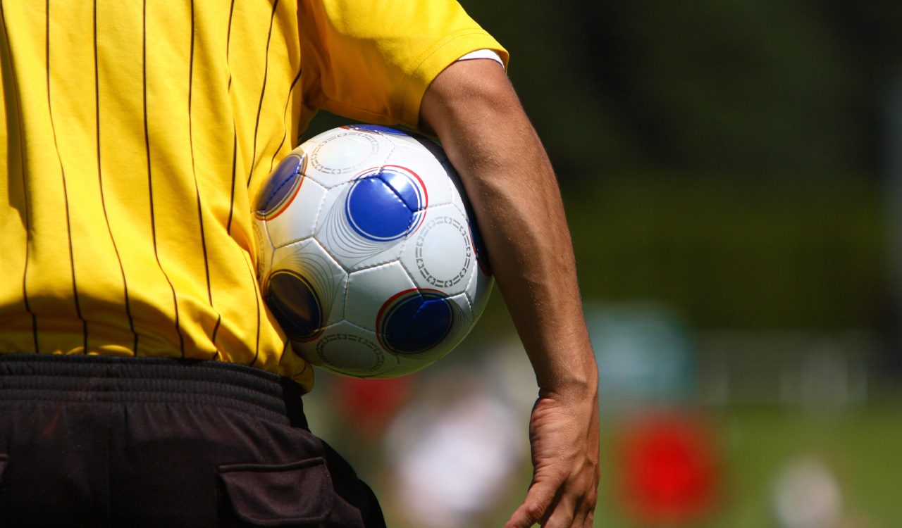 Person in yellow shirt holds a soccer ball and faces playing field.