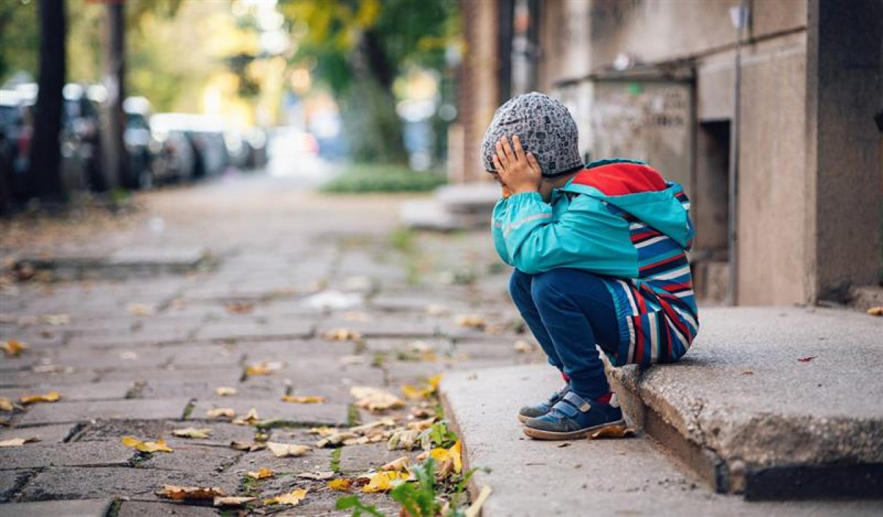 A young child in a wool hat sits on a stair outside a building on a city street.