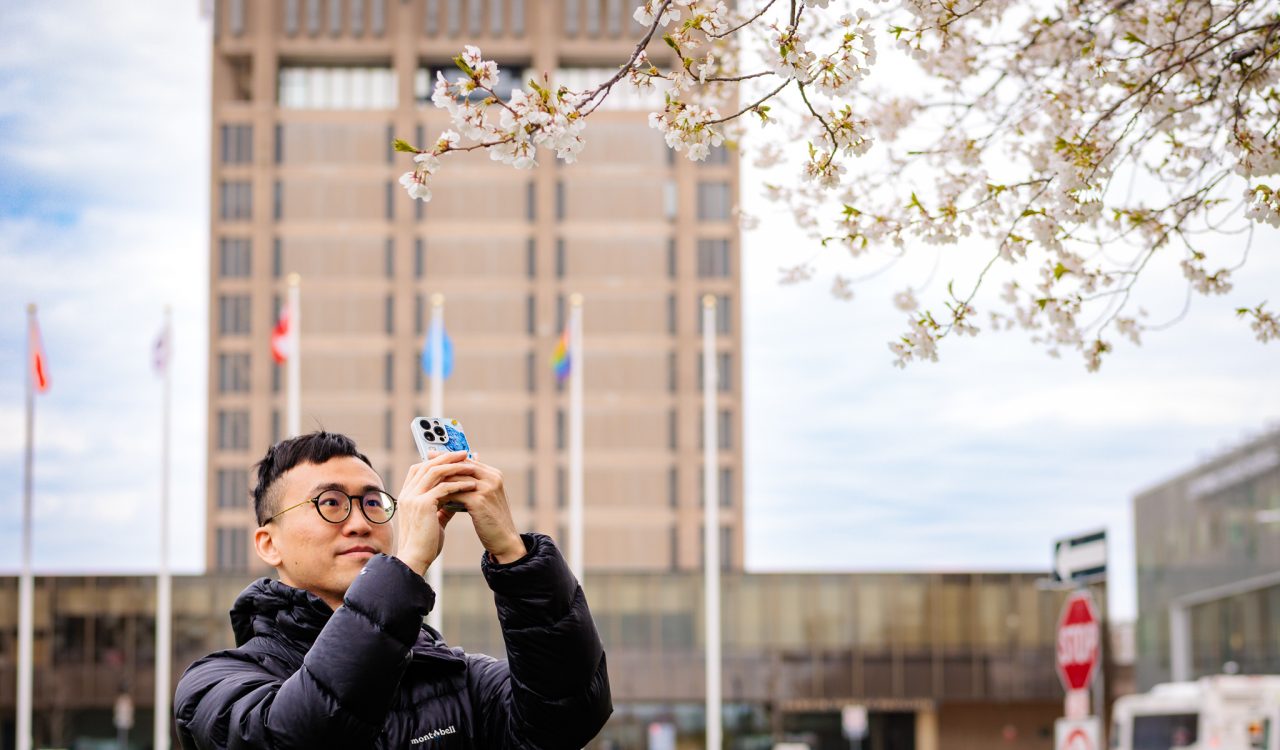 A person in a black puffer jacket and glasses stands outdoors beneath blooming cherry blossom branches, holding up a smartphone to take a photo. A tall building and several flagpoles are blurred in the background under a cloudy sky.