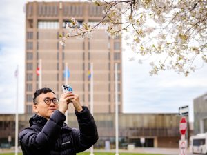 A person in a black puffer jacket and glasses stands outdoors beneath blooming cherry blossom branches, holding up a smartphone to take a photo. A tall building and several flagpoles are blurred in the background under a cloudy sky.