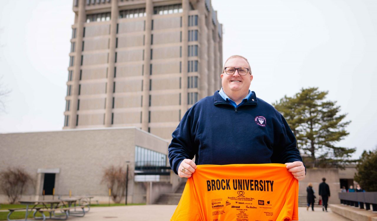 A man holds an orange shirt outside in front of a tall concrete tower.
