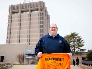 A man holds an orange shirt outside in front of a tall concrete tower.