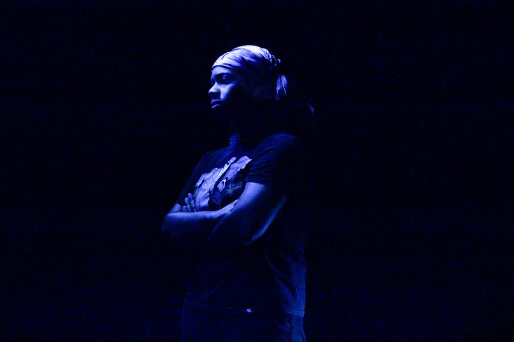 A performer stands on a stage under dark, blue light.