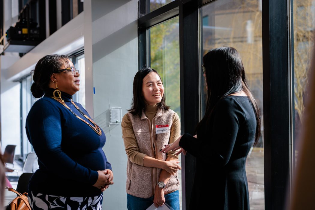 Three people have a conversation while standing beside a window.