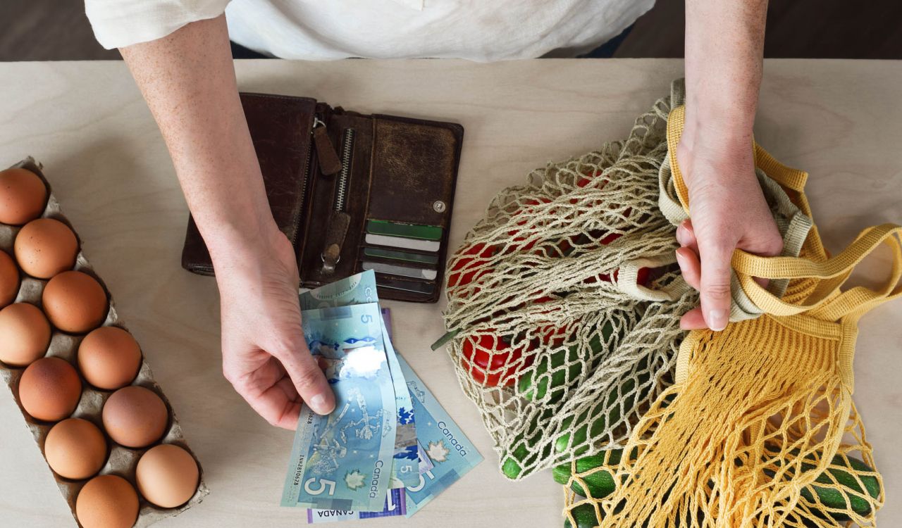 Hands, seen from above, hold Canadian cash above groceries on a counter.