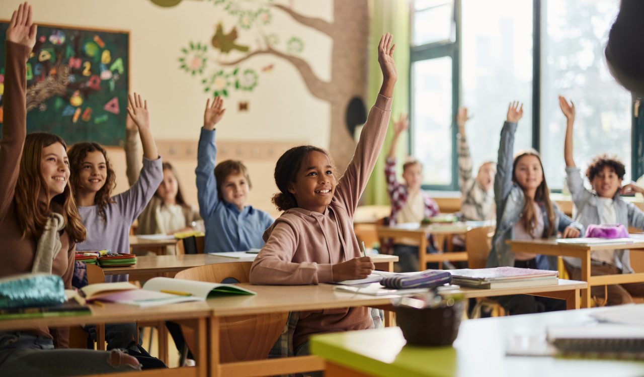 Large group of happy elementary students raise their hands in a classroom.