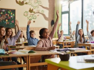 Large group of happy elementary students raise their hands in a classroom.