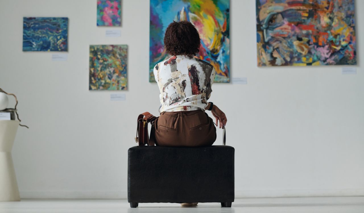 A woman sits on a bench looking at paintings in art gallery.