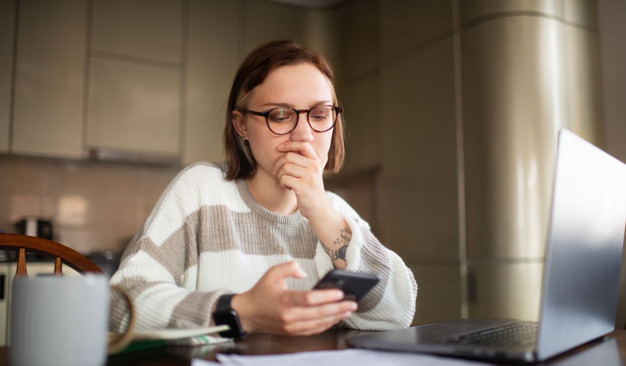 A young woman looks concerned while viewing content on phone and laptop.