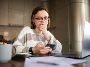 A young woman looks concerned while viewing content on phone and laptop.