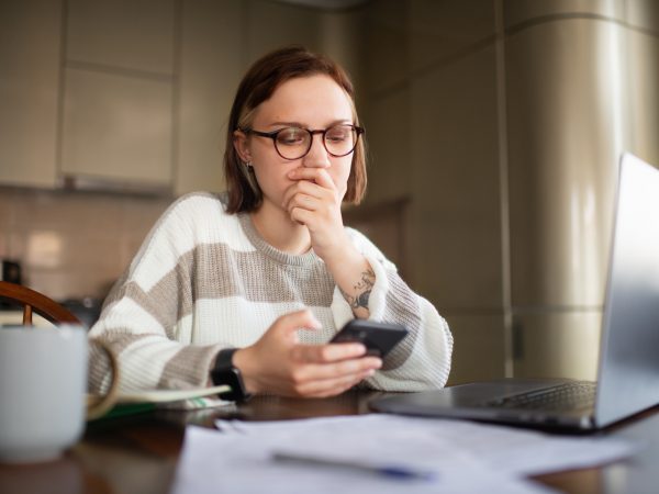 A young woman looks concerned while viewing content on phone and laptop.