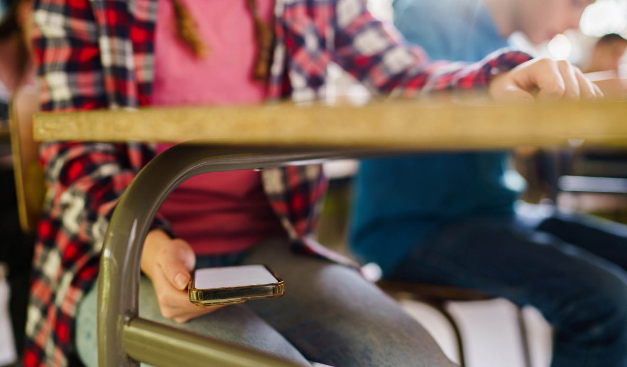A child holds a cellphone hidden under a desk in a classroom.