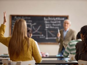 Teenage girl raising hand during classroom lesson while senior male teacher explaining graphs on chalkboard, showing active learning and student engagement.