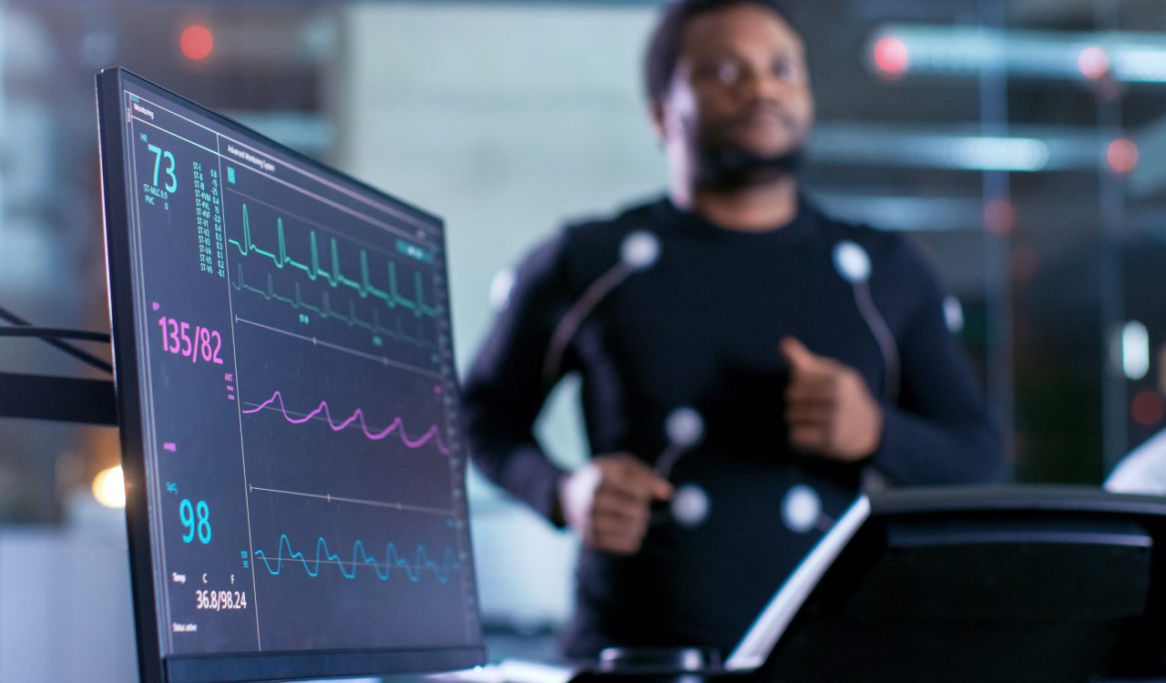 A close-up of a monitor showing health information with a person running on a treadmill behind it.