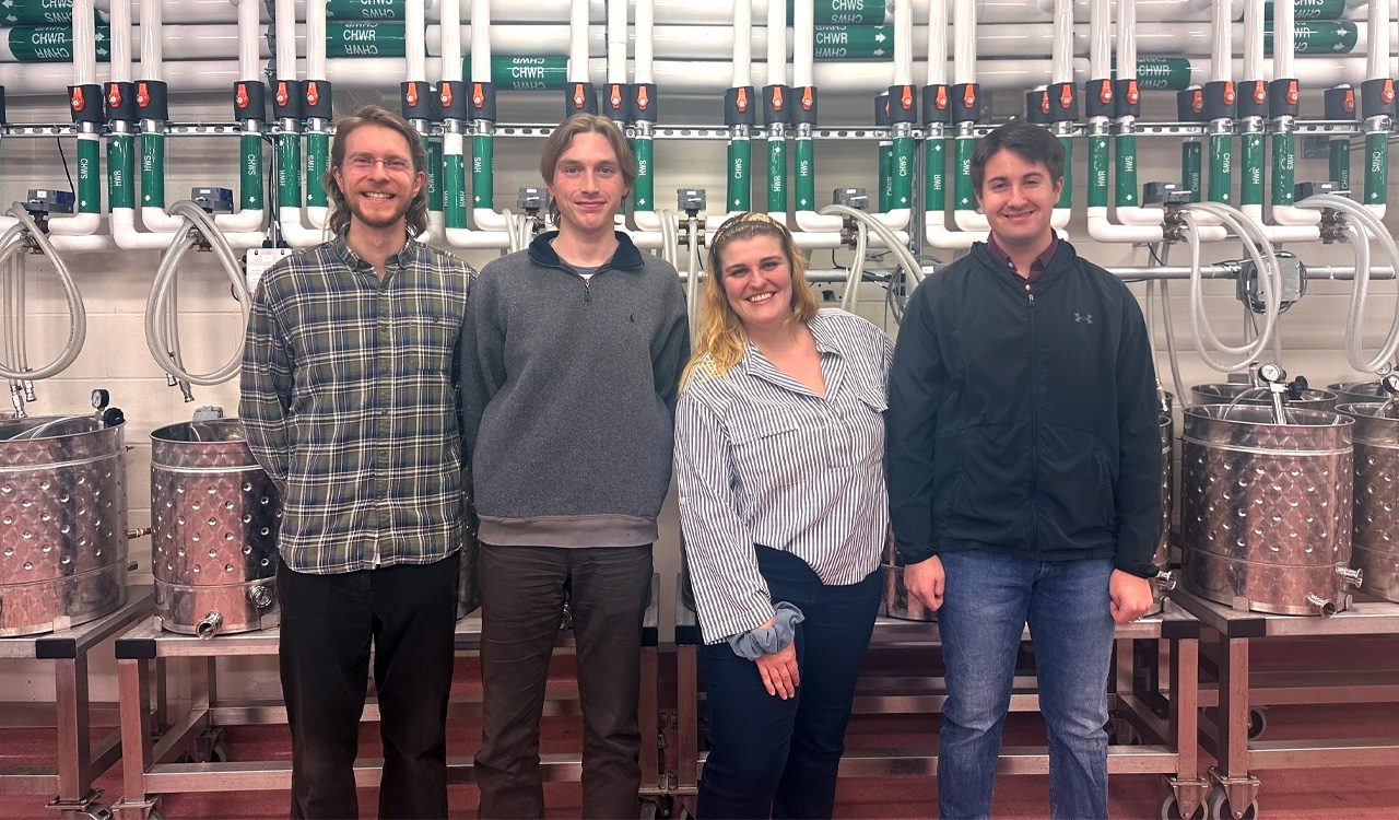 Four university students stand in a line in front of winery equipment.