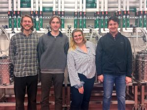 Four university students stand in a line in front of winery equipment.