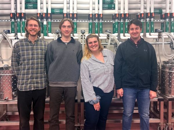 Four university students stand in a line in front of winery equipment.