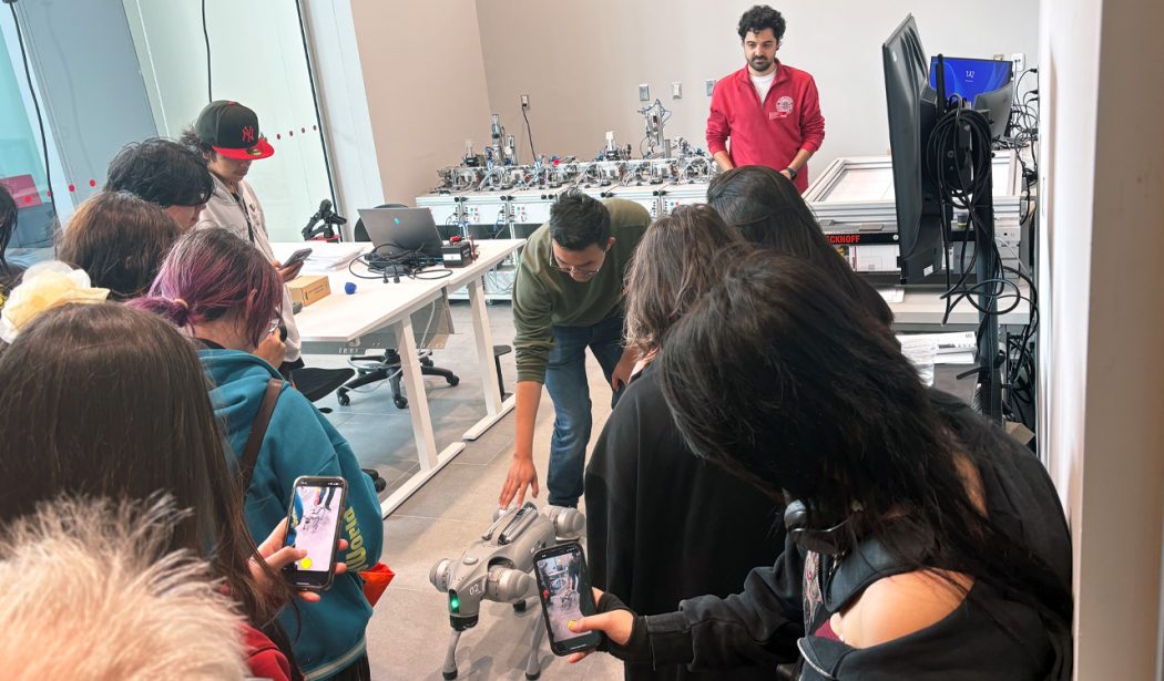 A group of high school students observe a robotic dog in a university classroom.