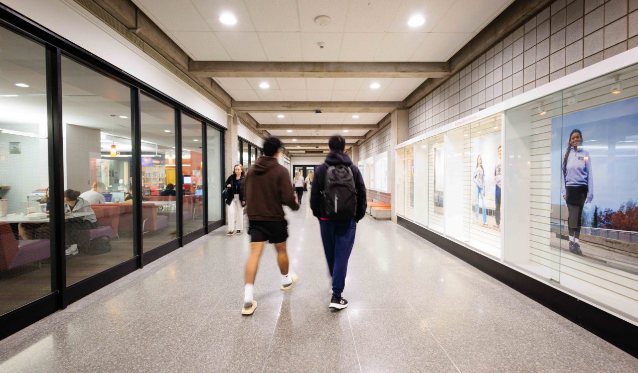 Two people walk down a brightly lit hallway with newly installed LED lighting at Brock University, with glass-walled study spaces on one side and illuminated display cases on the other.