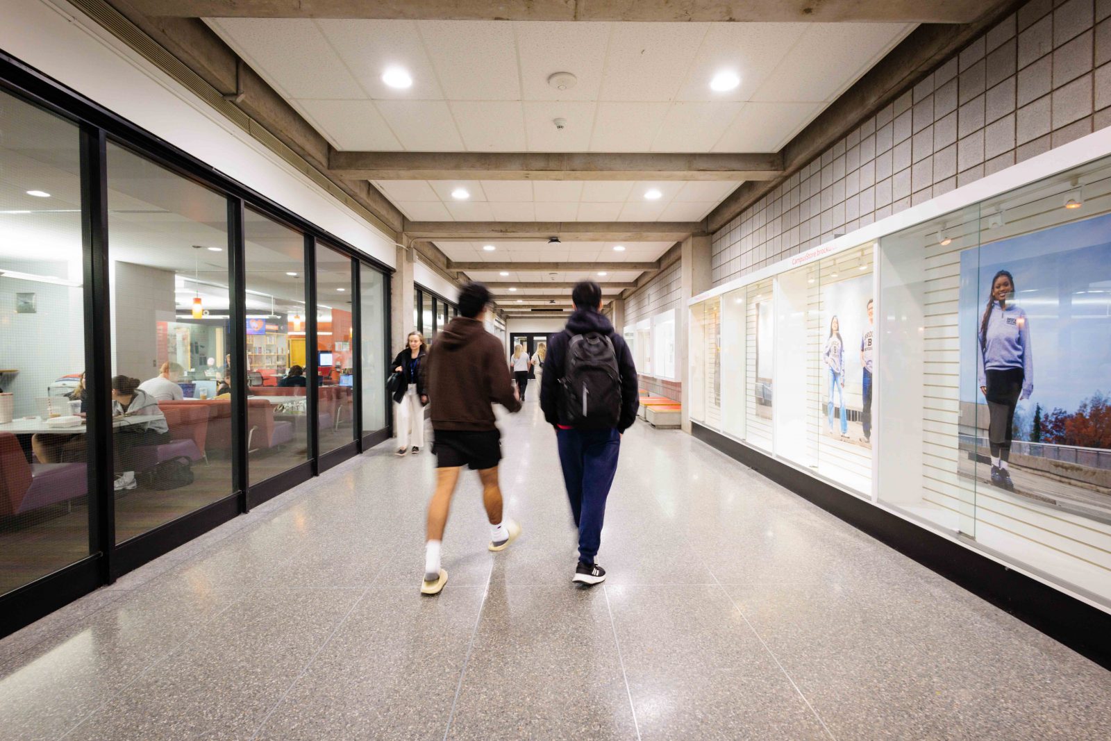 Two people walk down a brightly lit hallway with newly installed LED lighting at Brock University, with glass-walled study spaces on one side and illuminated display cases on the other.