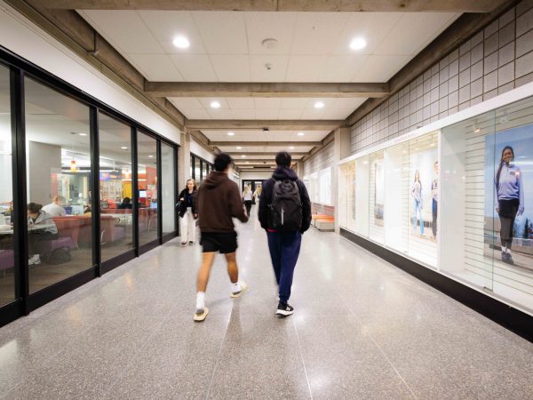 Two people walk down a brightly lit hallway with newly installed LED lighting at Brock University, with glass-walled study spaces on one side and illuminated display cases on the other.