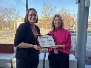 Two women pose with an award in front of a window.