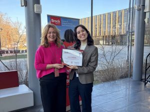 Two women pose with an award in front of a window.