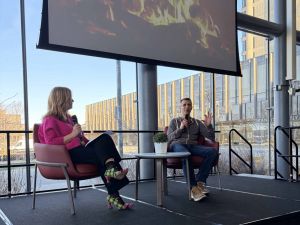 Two people sit on stage and have a conversation in front of a window.