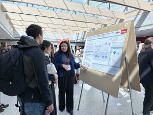 A young woman speaks to two other people about a research poster in a sunny room.