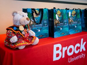 Bear wearing traditional Indigenous regalia rests on a table with gift bags.