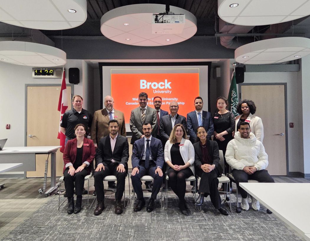 A large group of people pose in two rows in front of a presentation screen in a bright meeting room.