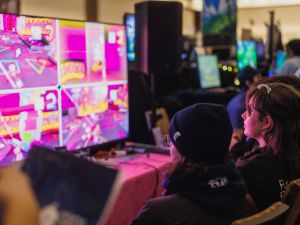 Two people sit in front of a large, colourful screen playing a video game.
